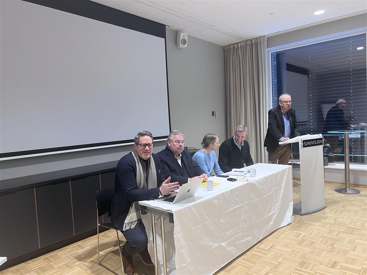 Mennesker ved et bord, et menneske på talerstol - Klikk for stort bilde