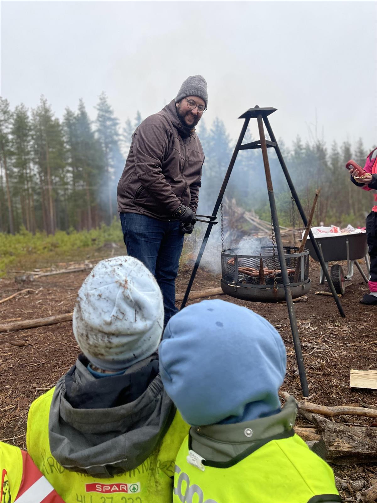 Mann som griller pølser, barn ser på - Klikk for stort bilde